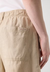 Béžové bavlněné kraťasy s elastickým pasem, které mají jedno zadní kapsu a malé červenobílé logo "Calvin Klein".