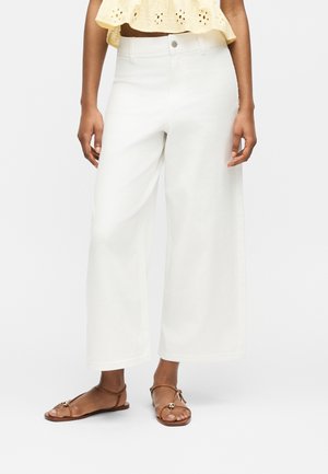 Persoon die witte, hooggetailleerde, wijde broek draagt, een gele cropped top en bruine sandalen met bandjes en platte zolen, staand tegen een effen achtergrond.