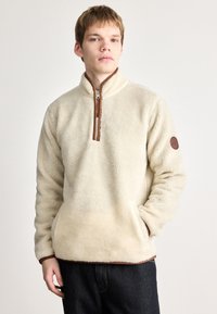 Cremfarvet fleece-pullover med høj krave, brun lynlås og læderdetalje på venstre ærme. Har en afslappet pasform og struktureret stof.