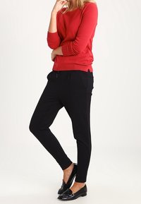 Pull en tricot rouge à manches longues associé à un pantalon noir fuselé. Mocassins en vernis noir et teint de peau clair visibles.