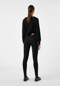 Suéter de punto negro, combinado con jeans negros y zapatos negros. Los jeans tienen un parche en la parte trasera y un diseño ajustado, acentuando la silueta.