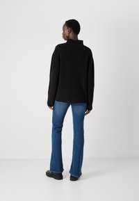 Svart ribbestrikket genser med høy krage, kombinert med blå slengjeans og svarte sko; vist fra baksiden.