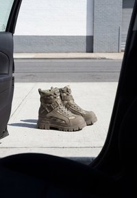 Paire de bottes de combat couleur beige placées sur un trottoir ensoleillé vues à travers la porte ouverte d’un véhicule stationné.