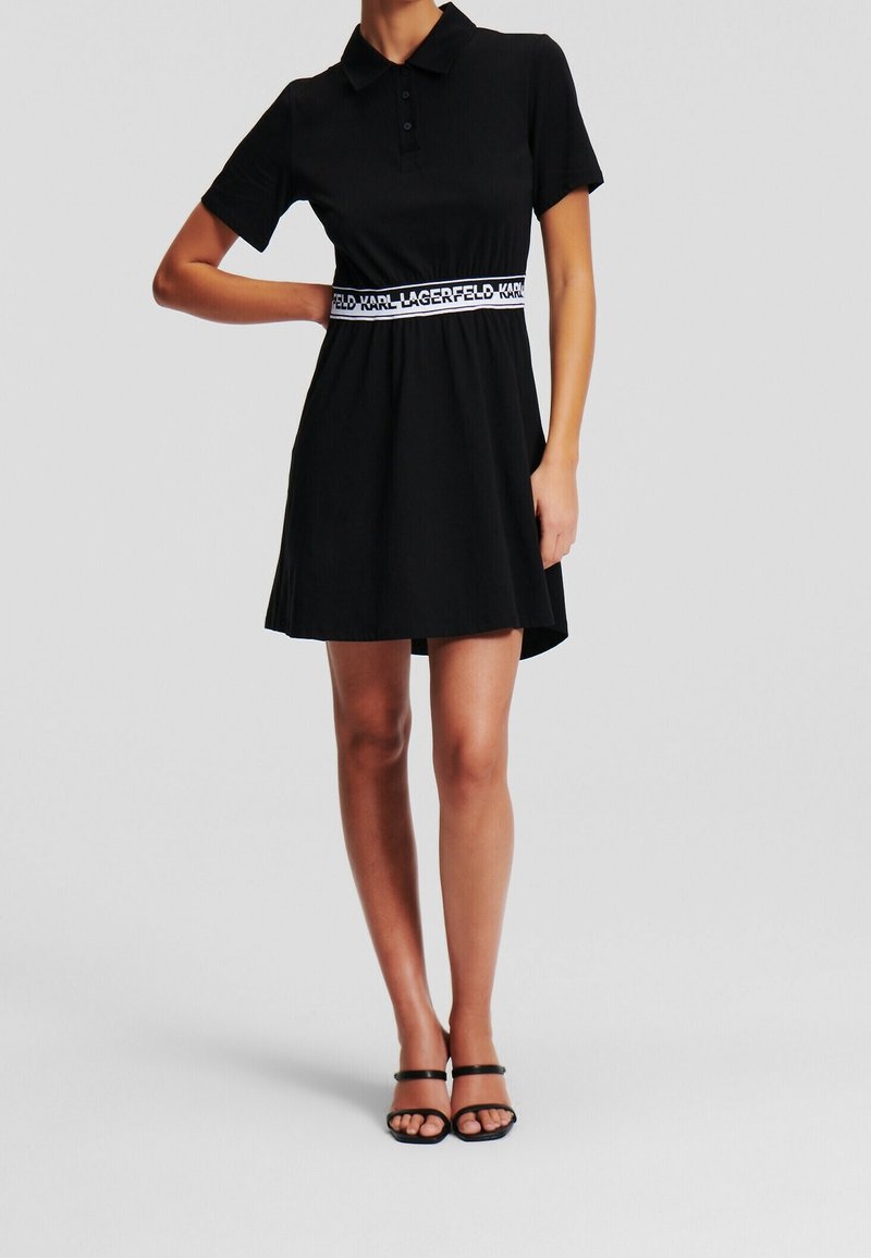 Svart polo-klänning med korta ärmar och figursydd midja, med ett kontrasterande märkes-elastiskt bälte. Slät tyg och utsvängd kjol.