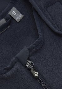 Mörkblå fleecejacka med detalj av dragkedjeslutning, mjuk textur och tygetikett. Dragkedjan har en logotypdekorerad dragtab och förstärkta kanter.
