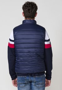 Chaleco acolchado azul marino con cuello alto, con secciones de acolchado horizontal. Lleva puesto sobre una camisa de manga larga multicolor y jeans de denim.