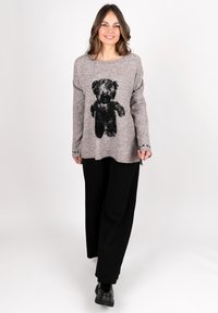 Beige gestrickter Pullover mit einem schwarzen Teddybär-Graphic auf der Vorderseite. Kombiniert mit schwarzen weit geschnittenen Hosen und schwarzen Stiefeletten.