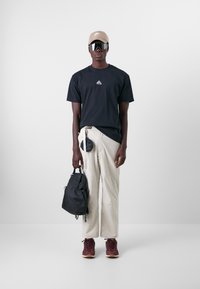 Schwarzes T-Shirt mit kleinem Logo, beiger Baseballcap, reflektierende Sonnenbrille, cremefarbene Hose, schwarzer Rucksack und kastanienbraune Sneakers mit Gummisohlen.