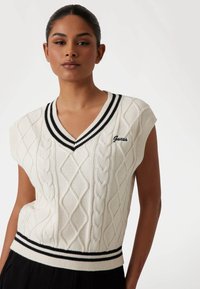 Knit sleeveless sweater in cream with cable pattern, V-neck, and black-striped ribbed hem. Features embroidered "Guess" logo in black.