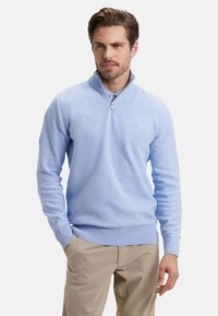 Sweat à zip bleu clair en tricot texturé ; avec un col montant et des poignets et un ourlet côtelés ; associé à un pantalon beige.