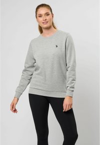 Grijze sweater van zacht materiaal, met lange mouwen, geribde boorden en onderrand, en een klein zwart logo op de borst. Gepaard met zwarte leggings.