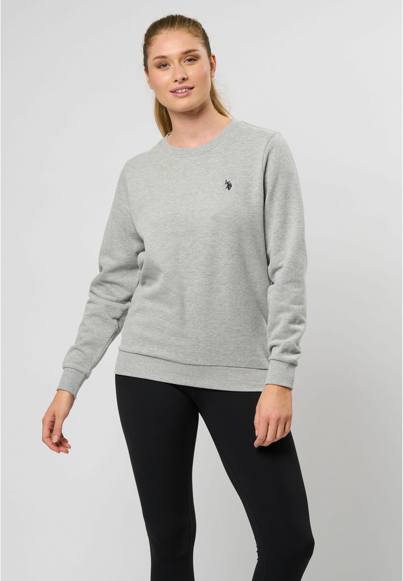 Grijze sweater van zacht materiaal, met lange mouwen, geribde boorden en onderrand, en een klein zwart logo op de borst. Gepaard met zwarte leggings.