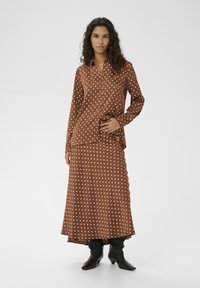 Braune Bluse mit weißen Punkten und Kragen, kombiniert mit einem passenden langen Rock. Beide Kleidungsstücke haben eine glatte Textur. Schwarzes Schuhwerk rundet den Look ab.