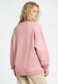 Femme blonde portant un sweat-shirt rose ample et un pantalon beige, légèrement tournée avec une expression neutre sur un fond clair uni.