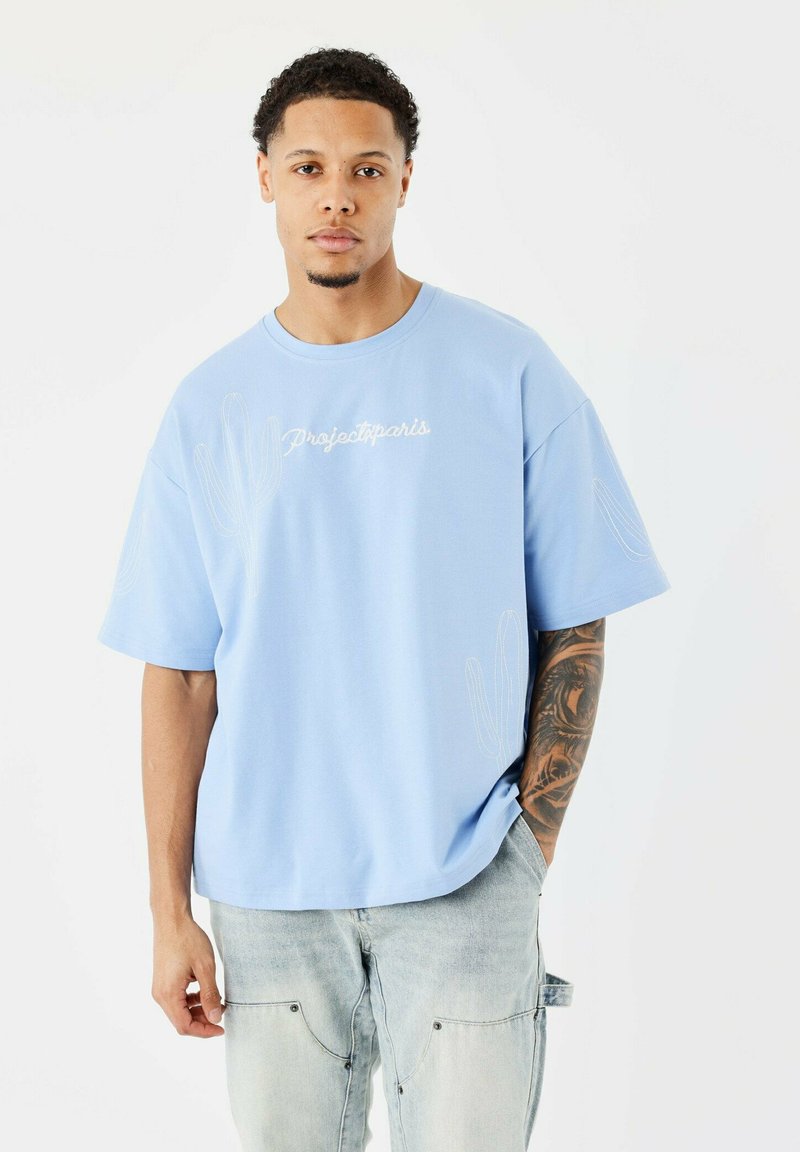 Camiseta de manga corta azul claro con un corte relajado, que presenta diseños de cactus bordados en blanco y el texto "Projectparis" en la parte frontal.