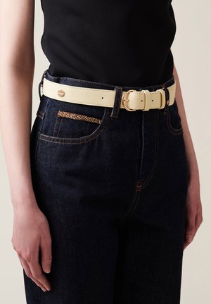 Cremefarbener Kunstledergürtel mit zwei metallischen Schnallen, getragen über dunkelblauen Jeans mit orangefarbener Naht und einem kleinen gewebten Taschendetail.