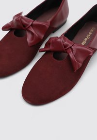 Bailarinas burdeos con un acabado de ante suave, que cuentan con un prominente lazo de cuero y un diseño de punta redonda.