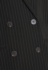 Svart randig kavaj i texturerad tyg, med fyra runda bruna knappar och slagkrage. Har sidofickor.