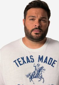 T-shirt blanc à manches courtes présentant un texte imprimé bleu "TEXAS MADE" et un graphique d'un cow-boy à cheval. Le tissu semble doux et léger.