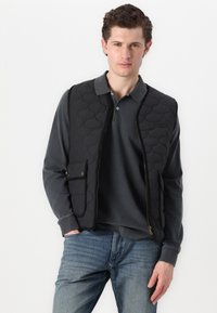 Melnā kvadrātveida veste ar pūkaina tekstūru, ar rāvējslēdzēju un sānu kabatu. Mugurā valkā pelēku garu piedurkņu polo un pievienotas zilas džinsus.