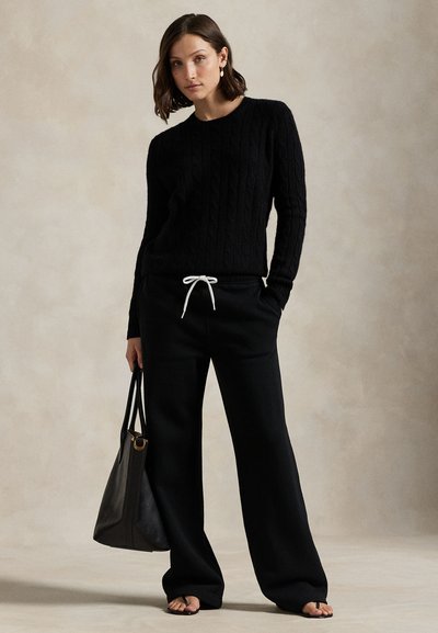 Kvinde iført sort kabelstrikket sweater, sorte vidde bukser med hvid snor, sorte sandaler, der holder en stor sort taske, står mod ensfarvet baggrund.