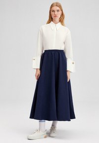Bílá blůza s límcem a oversized rukávy, doplněná knoflíkovými detaily, v kombinaci s dlouhou námořnickou sukní, a bílými teniskami a ponožkami.