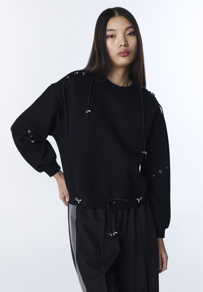 Jeune femme portant un sweat-shirt noir avec des œillets métalliques et des cordons, posant devant un fond blanc uni.