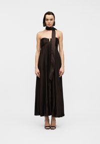 Γυναίκα που φοράει αμάνικο, πλισέ μακρύ φόρεμα στο σκούρο καφέ χρώμα με ένα μακρύ κασκόλ στο ίδιο χρώμα, στέκεται όρθια μπροστά σε απλό λευκό φόντο.