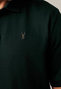 Ciemnozielona koszulka polo z teksturowanej tkaniny, z małym, haftowanym logo jelenia na lewej stronie klatki piersiowej.