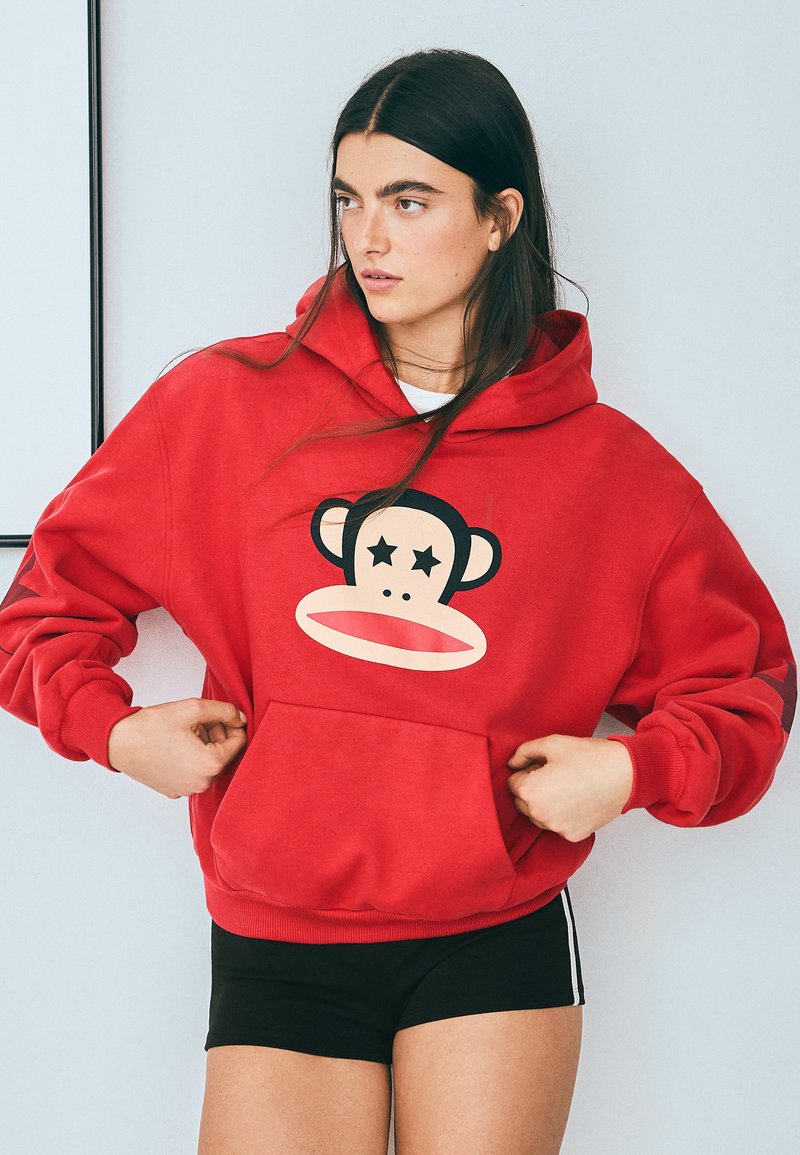 Jeune femme aux longs cheveux foncés portant un sweat à capuche rouge avec un motif visage de singe et un short noir, posant les mains ajustant le sweat à capuche.