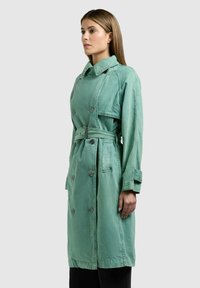 Nő hosszú, dupla soros, zöld ballonkabátban, övvel, oldalnézetben áll, egyszínű háttér előtt.