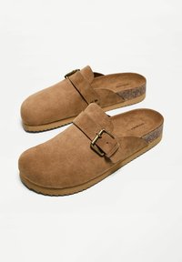 Rjavi semiš natikači s širokim trakom in nastavljivo kovinsko zaponko, z pluto podplatom in teksturiranim gumijastim podplatom. Gladek notranji del.