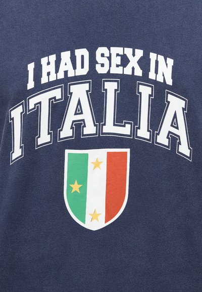 Μπλε μπλουζάκι ναυτικού με τολμηρό λευκό κείμενο: «ΚΑΝΑ ΝΤΟΥΣ ΣΤΗΝ ΙΤΑΛΙΑ» και γραφικό θυρεό με κάθετες ρίγες σε πράσινο, λευκό και κόκκινο και αστέρια.