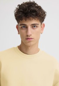 Sweat-shirt jaune clair avec un col rond, une texture lisse et une coupe décontractée. Le modèle a les cheveux courts et bouclés et porte une petite boucle d'oreille.