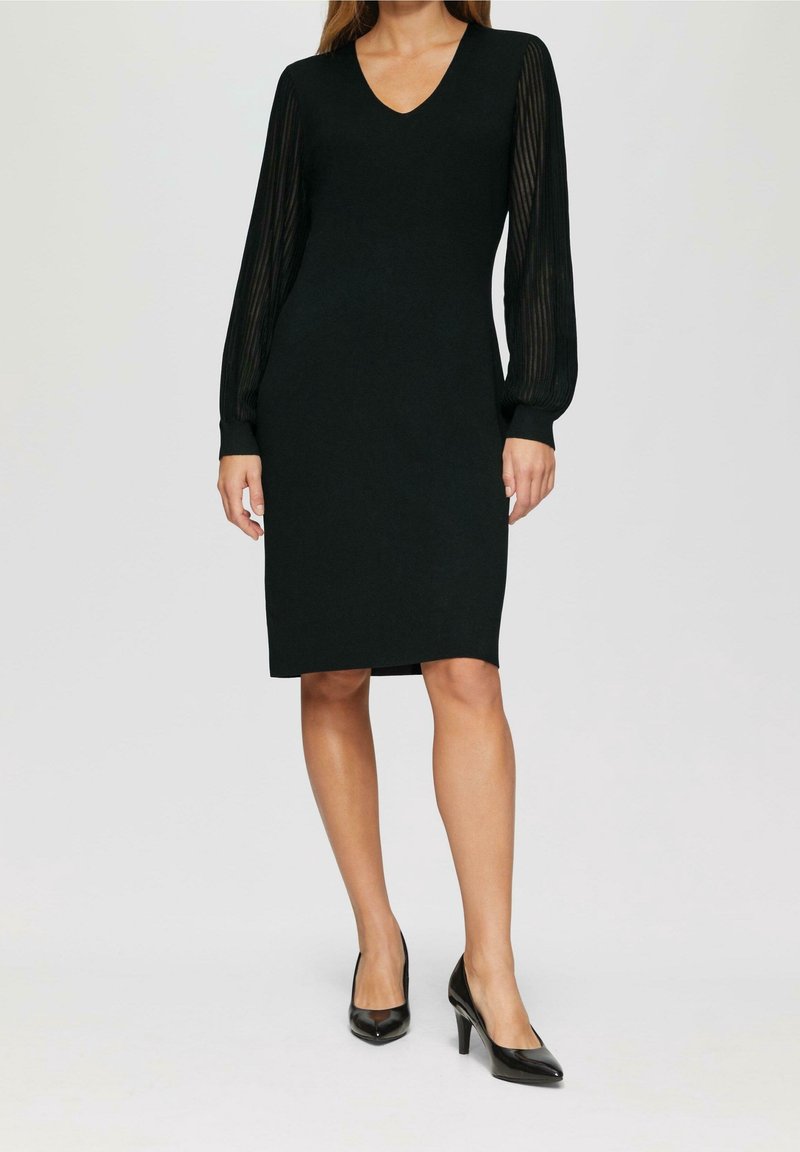 Robe noire avec un décolleté en V, des manches longues en tulle rayé, et une silhouette ajustée, associée à des chaussures à talons hauts noires.