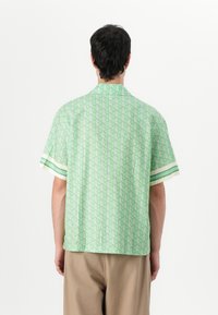 Green short-sleeve shirt with geometric pattern, featuring a collared neck and striped accents on sleeves. Lightweight fabric, relaxed fit.