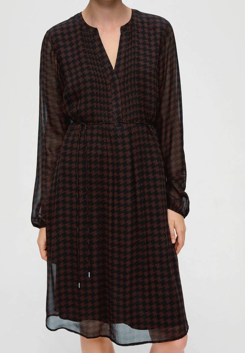 Femme portant une robe noire à manches longues avec des motifs géométriques marron et un col en V, avec une taille cintrée et des manches en tissu transparent.