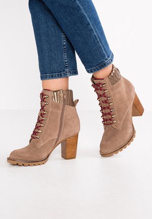 Personne portant des bottines en suède beige à lacets avec talons blocs en bois et jean en denim bleu sur fond blanc.
