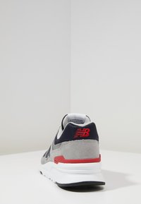 Baskets grises et bleu marine en daim et maille, avec une semelle blanche et une touche de rouge au niveau du talon, et un logo à l'arrière.