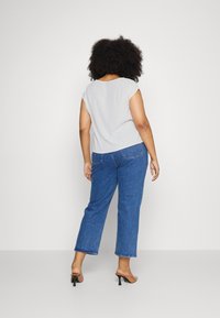 Kvinde med krøllet hår iført en hvid kortærmet top, blå jeans og sorte hælede sandaler, stående med ryggen til mod en enkel baggrund.