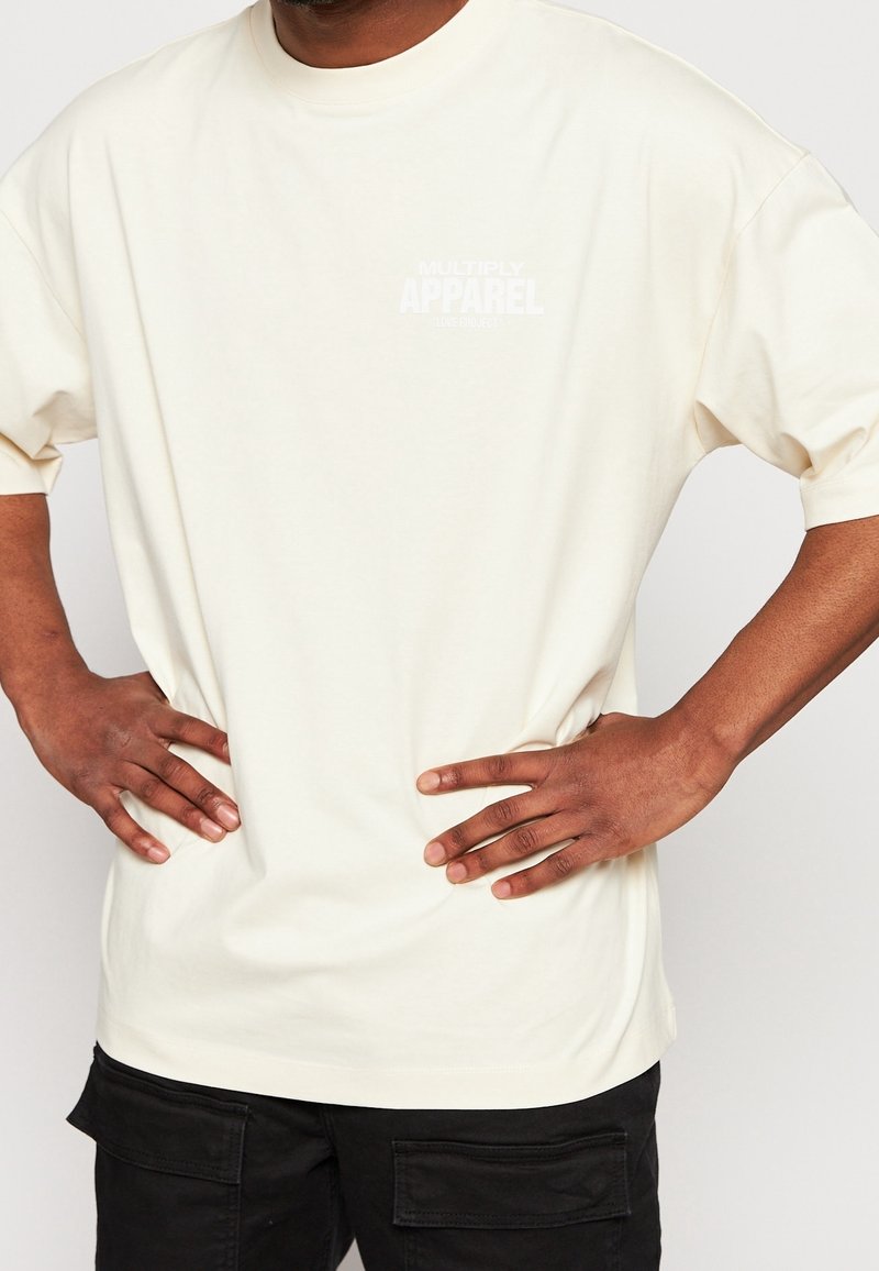 T-shirt en coton de couleur crème à manches courtes, arborant un petit logo blanc sur la poitrine avec l'inscription "MULTIPLY APPAREL".