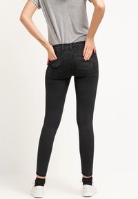 Jeans skinny negros de cintura alta, con dos bolsillos traseros y una textura de denim suave. El modelo lleva una camiseta gris y zapatillas blancas.