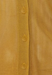 Gult stickat tyg med ett texturerat nätmönster, med fyra matchande runda knappar längs placket.