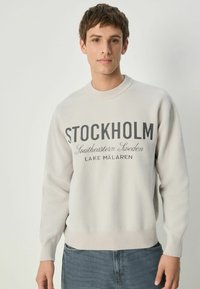 Lys grå sweatshirt med ribbet halsudskæring og manchetter. Har "STOCKHOLM" trykt tydeligt, og mindre tekst nedenfor. Blød tekstur.