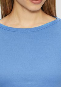Helles, blaues Baumwollhemd mit glatter Textur und weitem Ausschnitt; es zeichnet sich durch ein einfaches, unauffälliges Design ohne Muster oder Akzente aus.