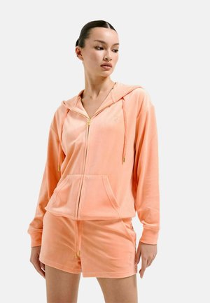 Femme portant un sweat à capuche zippé en velours couleur pêche et un short assorti, debout devant un fond gris clair uni.