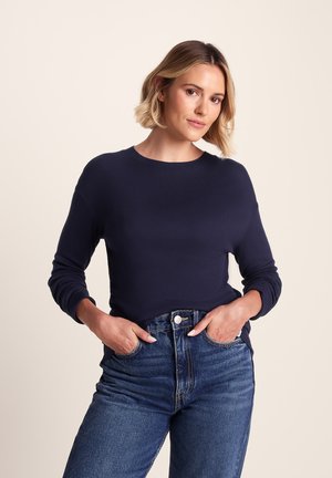Woman with short blonde hair wearing a navy long-sleeve shirt tucked into high-waisted blue jeans with hands in pockets.
