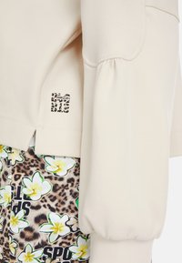 Beige Sweatshirt mit Puffärmeln, ausgestattet mit einem Leopardenmuster-Logoschild. Kombiniert mit gemusterten Shorts, die weiße Blumen und auffälligen Text zeigen.