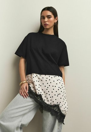 Young woman wearing a black oversized T-shirt layered over a polka dot skirt with lace trim and light washed wide-leg jeans, standing against a wall.