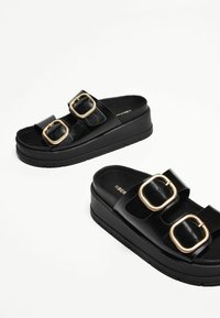 Sandales à plateforme noires avec deux brides noires brillantes et de grandes boucles dorées. Semelle intérieure texturée avec une finition lisse et une épaisse semelle noire.